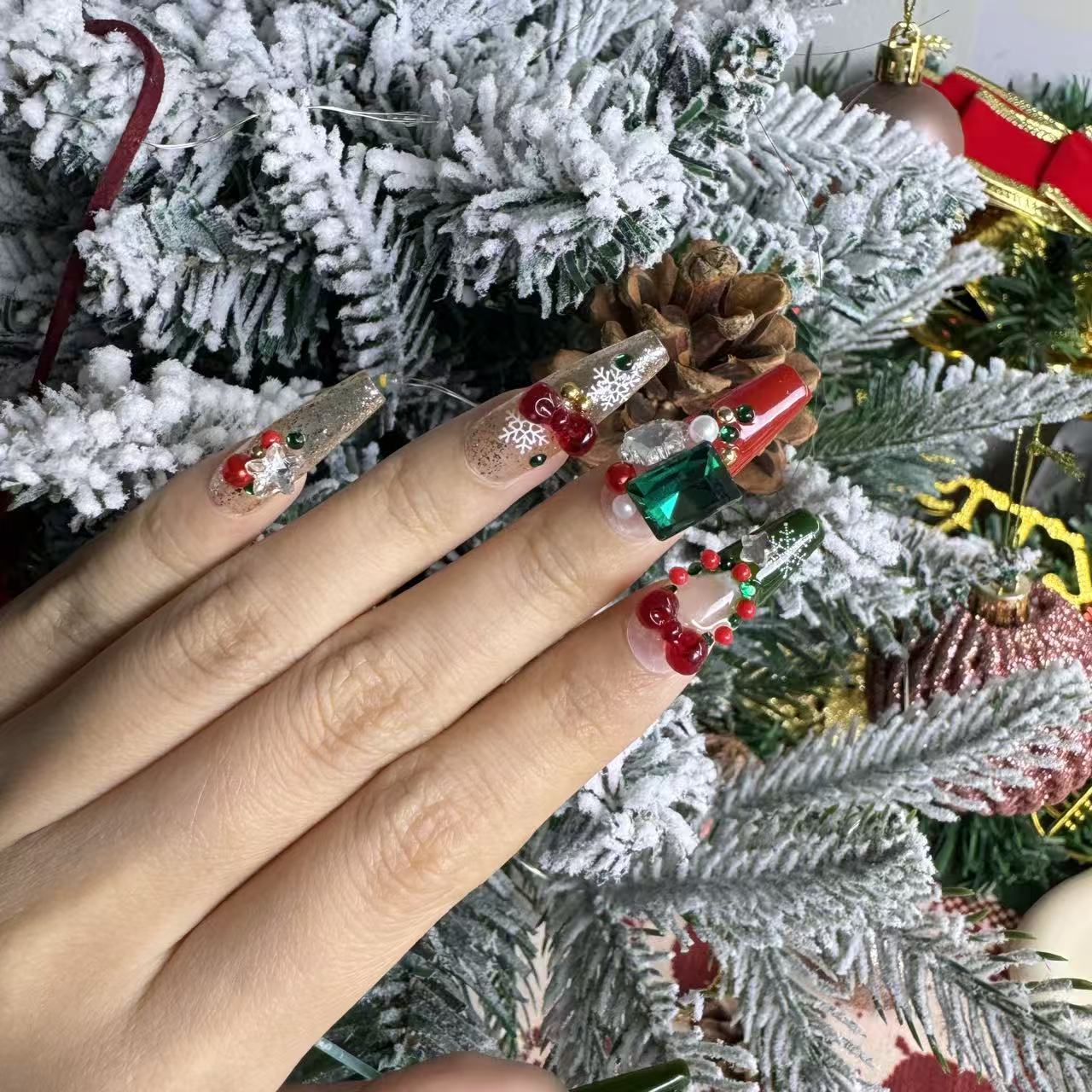 Festive Christmas nails in red, green and gold with large gems, ornament beads and 3D bell decorations, shown against snowy pine branches.