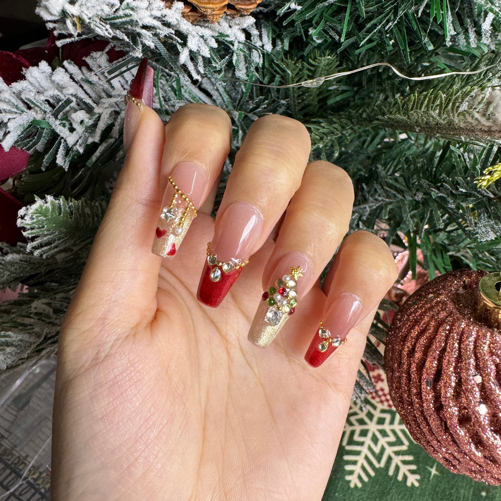 Hand wearing 3D Christmas tree nails with red glitter tips, gold accents and jeweled decorations, styled against snowy holiday greenery.