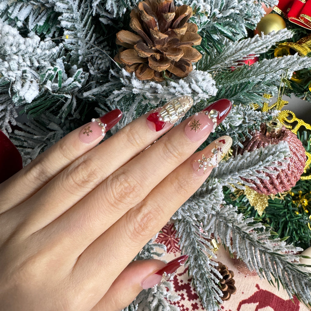 Hand wearing cute red and pink Christmas nails with 3D tree charms, glitter details and holiday accents against snowy pine branches.