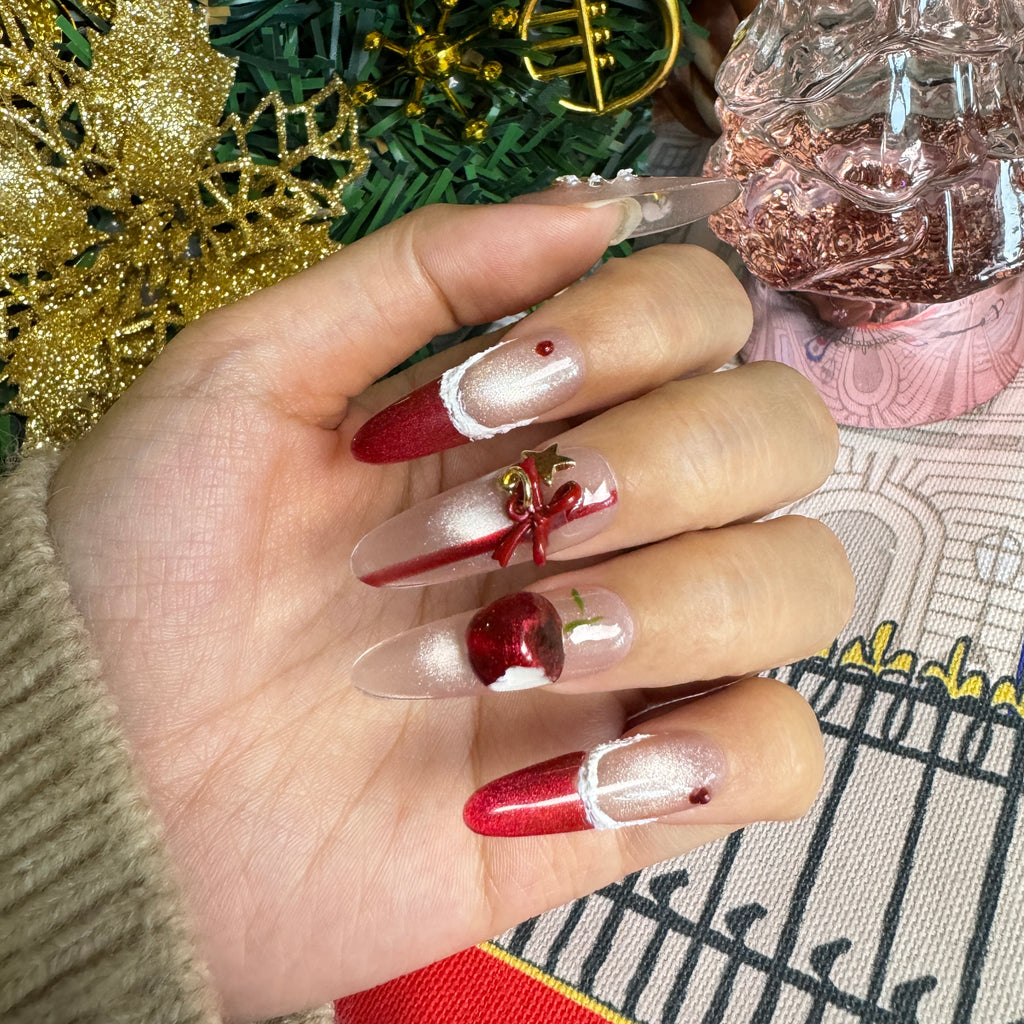 Close-up of festive red Christmas nails featuring 3D cherry charms, gold star accents and shimmer nude bases shown beside frosted Christmas greenery.