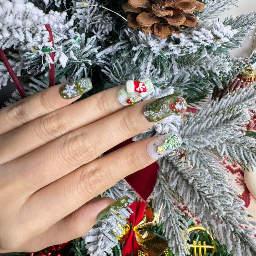 Hand wearing green Christmas nails featuring a 3D snow-globe charm, snowflakes and holiday tree details against festive pine branches.