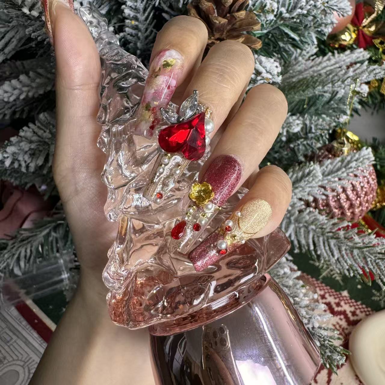 Close-up of red and gold Christmas press on nails featuring a bold ruby heart jewel, pearls and gold ornaments, styled with a pink crystal Christmas tree.