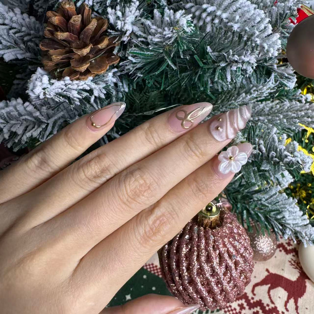 Hand wearing blush nude candy-themed press on nails with gold outlines, 3D pearly flowers and frosted swirl accents, posed against a Christmas tree background.