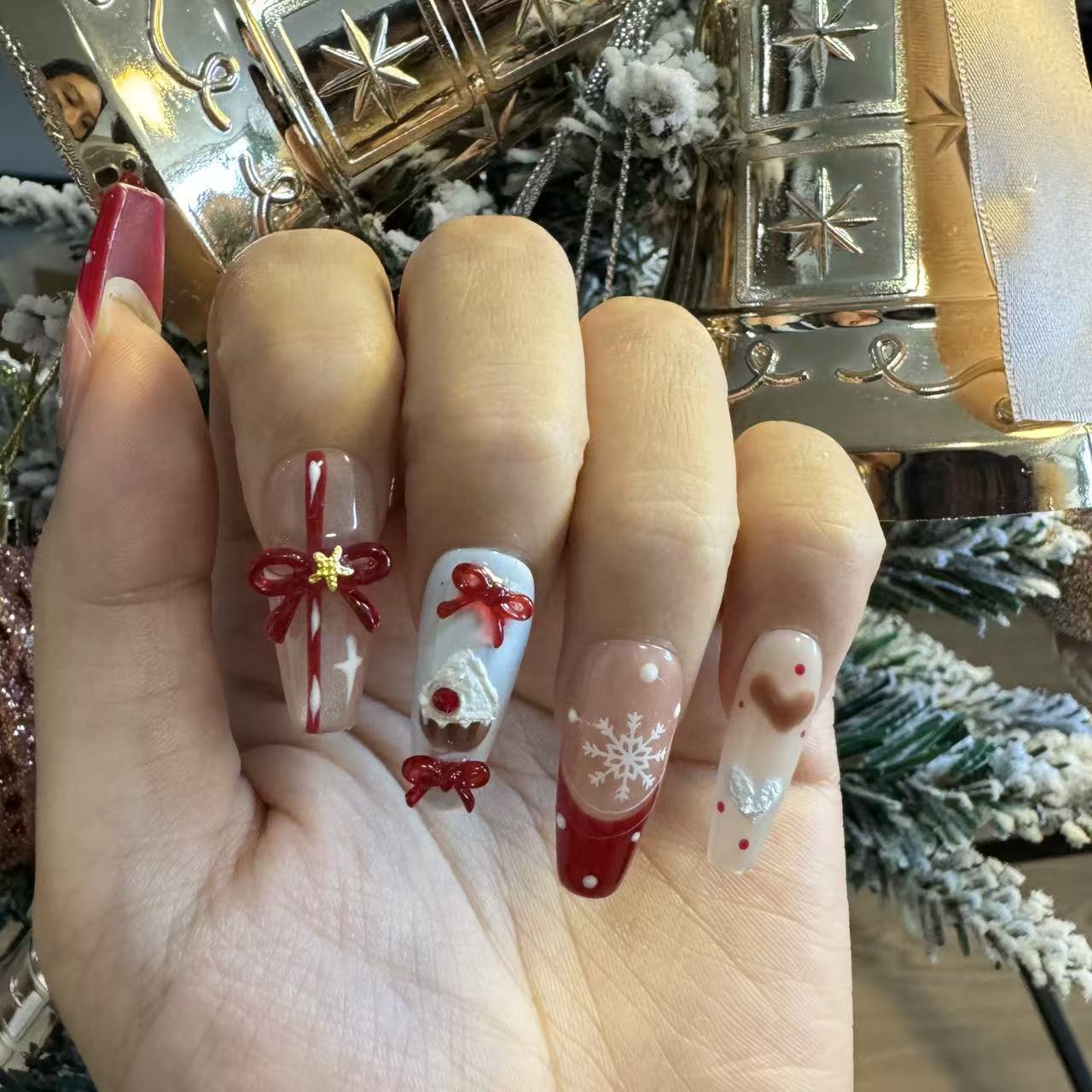 Close-up of Christmas press on nails featuring candy stripes, bows, red accents and winter snowflake details by a Christmas tree.