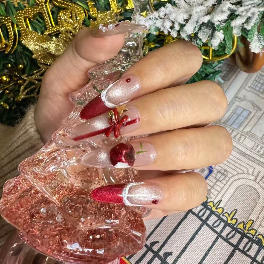 Hand wearing red and nude Christmas press on nails with 3D cherry ornaments, gold bows and white trim details, posed against a holiday tree background.