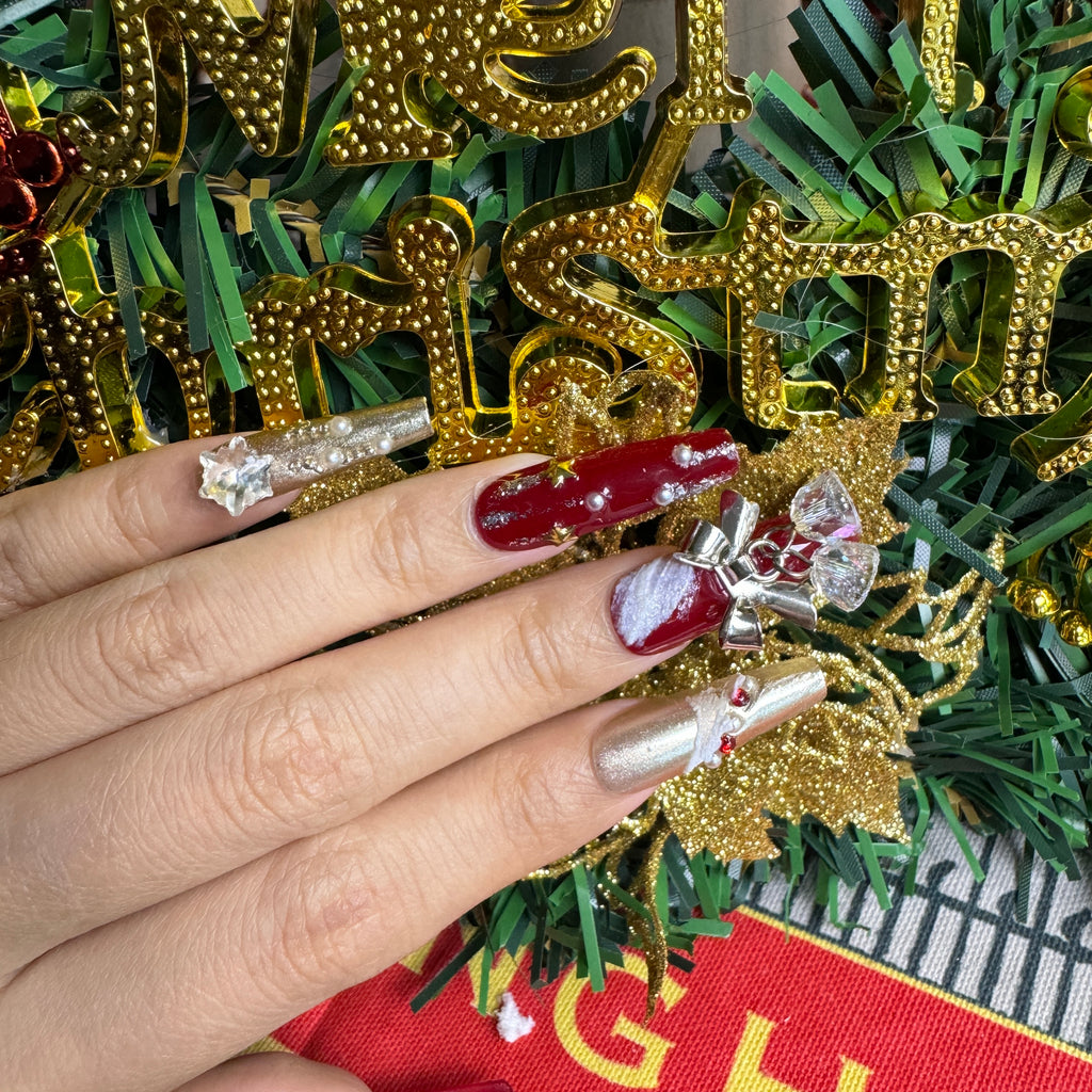 Hand wearing red Christmas nails with 3D bell charms, snowflake designs and gold details, styled with festive holiday decorations.