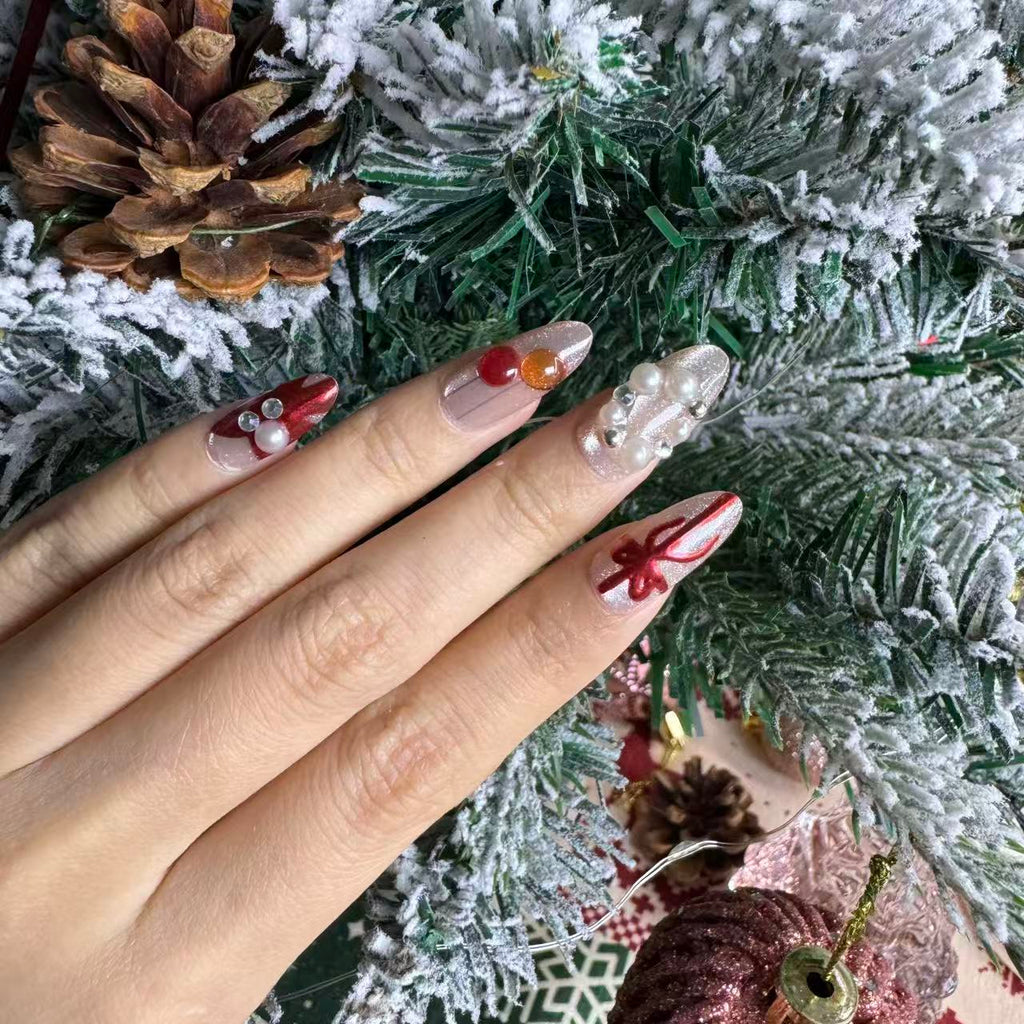 Hand wearing cute Christmas nails with red ribbons, pearls and soft pink shimmer, styled against snowy pine branches.