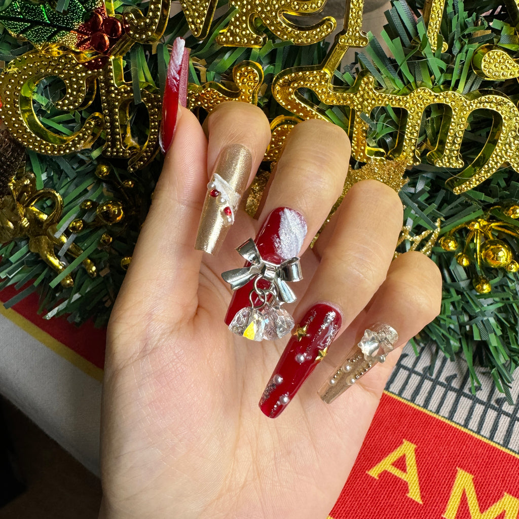Close-up of 3D Christmas nails featuring a crystal bell charm, textured snow accents and glossy red polish against snowy pine branches.