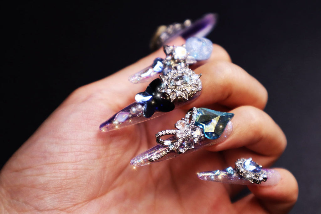 Close-up of luxury crystal press-on nails with butterfly design, pearl accents, and gemstone-inspired details on gradient purple base.