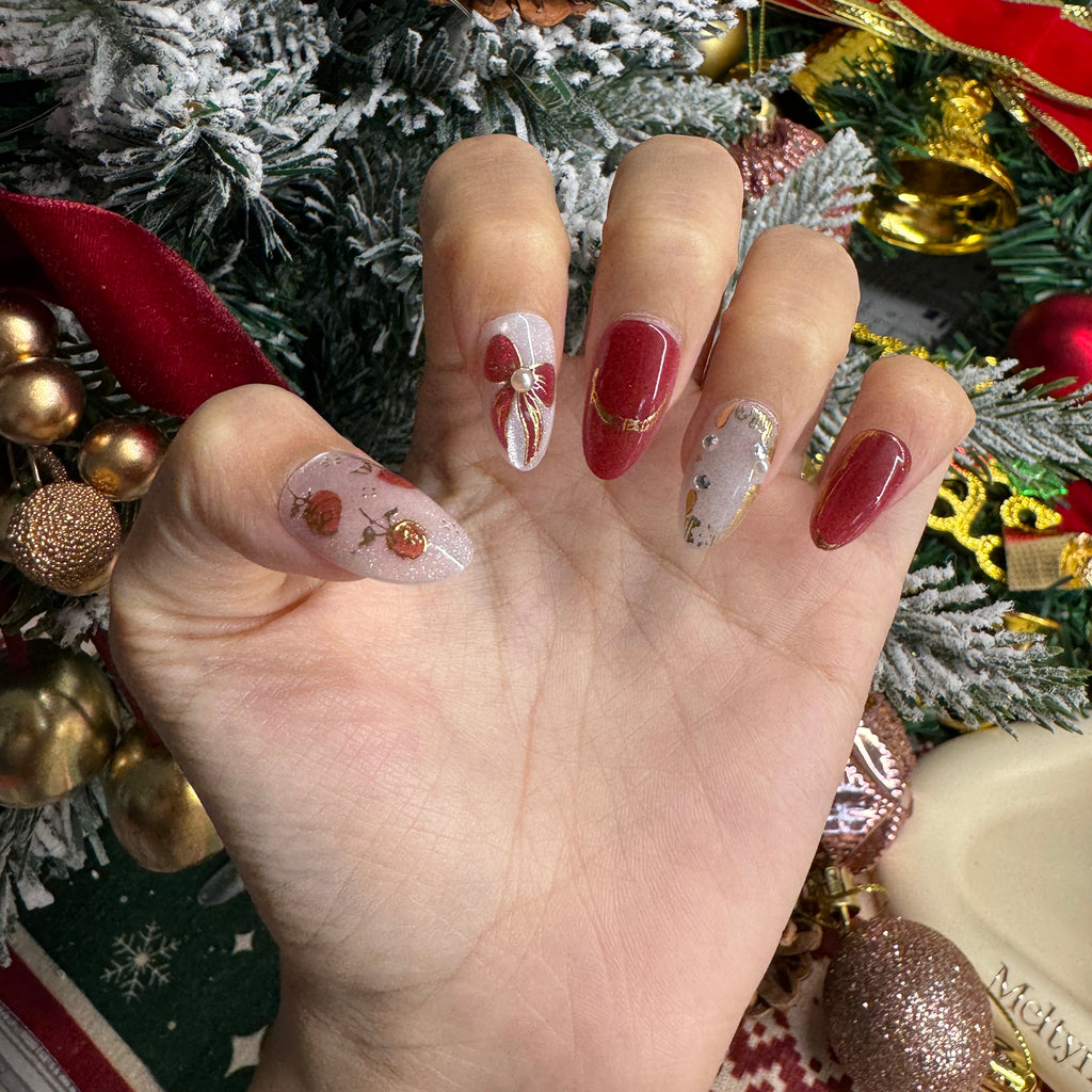 Hand wearing red and pearl-white Christmas nails with gold rose art and delicate ribbon accents, styled in a festive holiday setting.