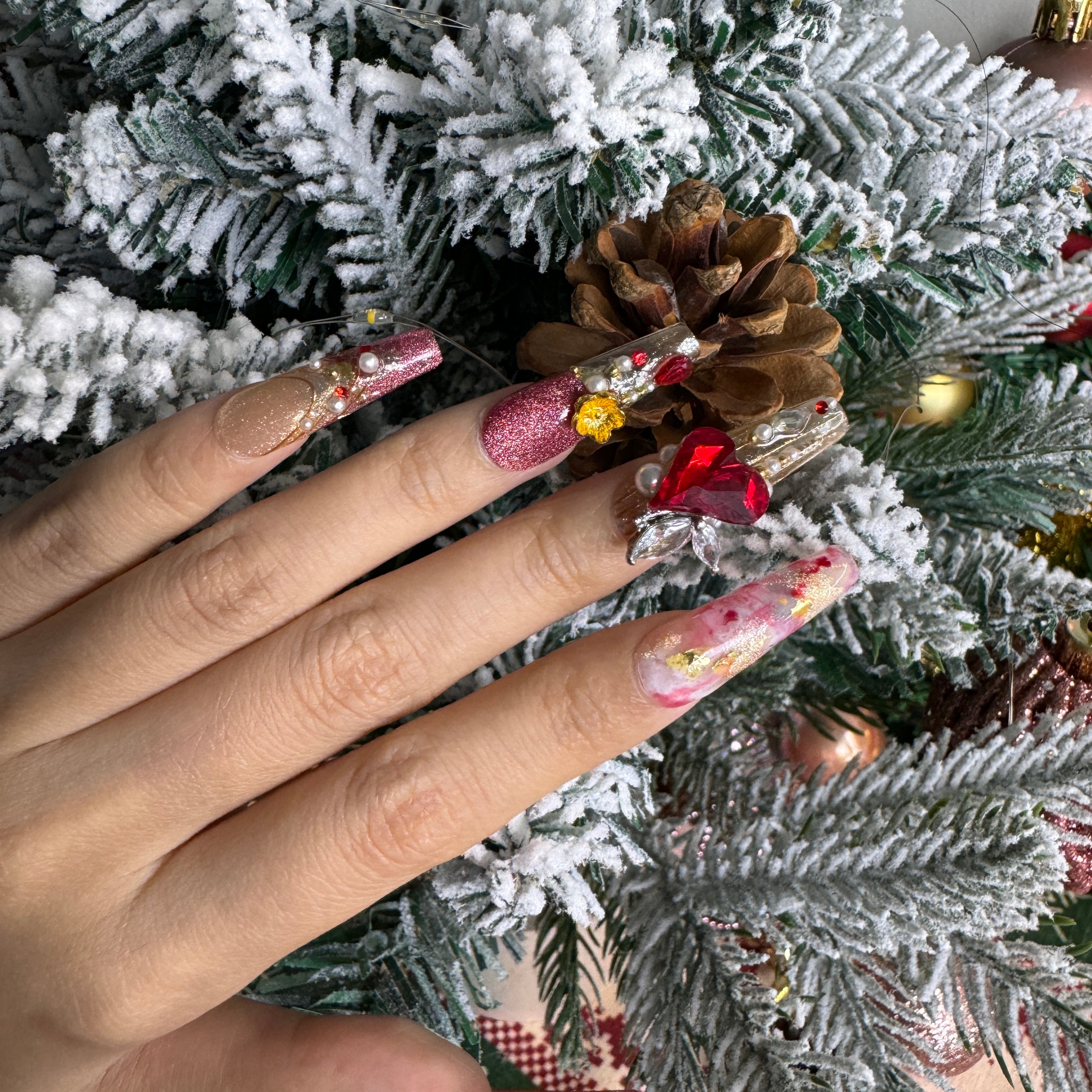 Red and gold luxury Christmas nails with amber gemstone, baroque patterns and pearl clusters, photographed against snowy holiday greenery.
