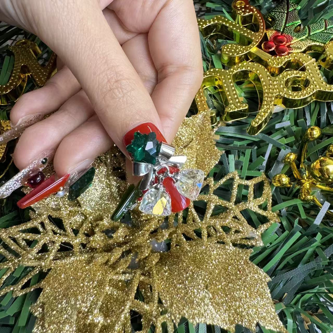 Close-up of Christmas press on nails with oversized green gems, silver bells and red accents, holding a gold holiday floral ornament.