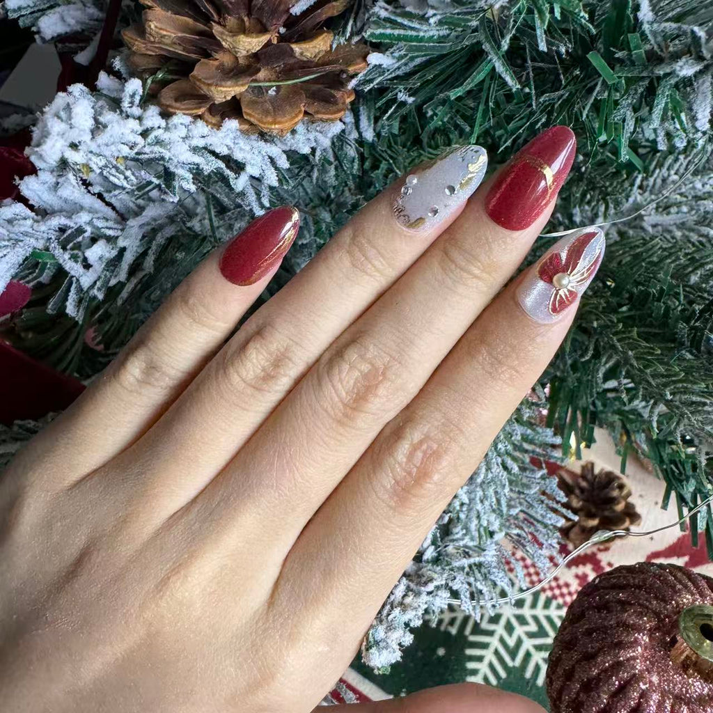 Red and ivory Christmas nails with gold floral detailing and subtle shimmer, photographed against snowy pine branches.