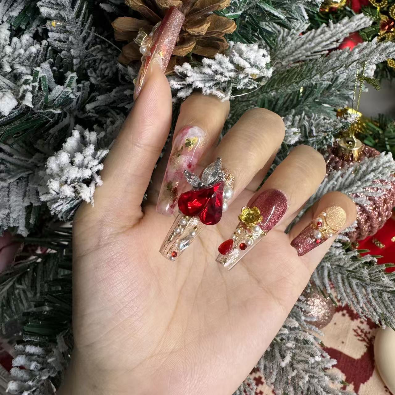 Hand wearing luxury red Christmas nails with a large ruby heart gemstone, gold foil accents and handcrafted baroque details beside a snowy Christmas tree.