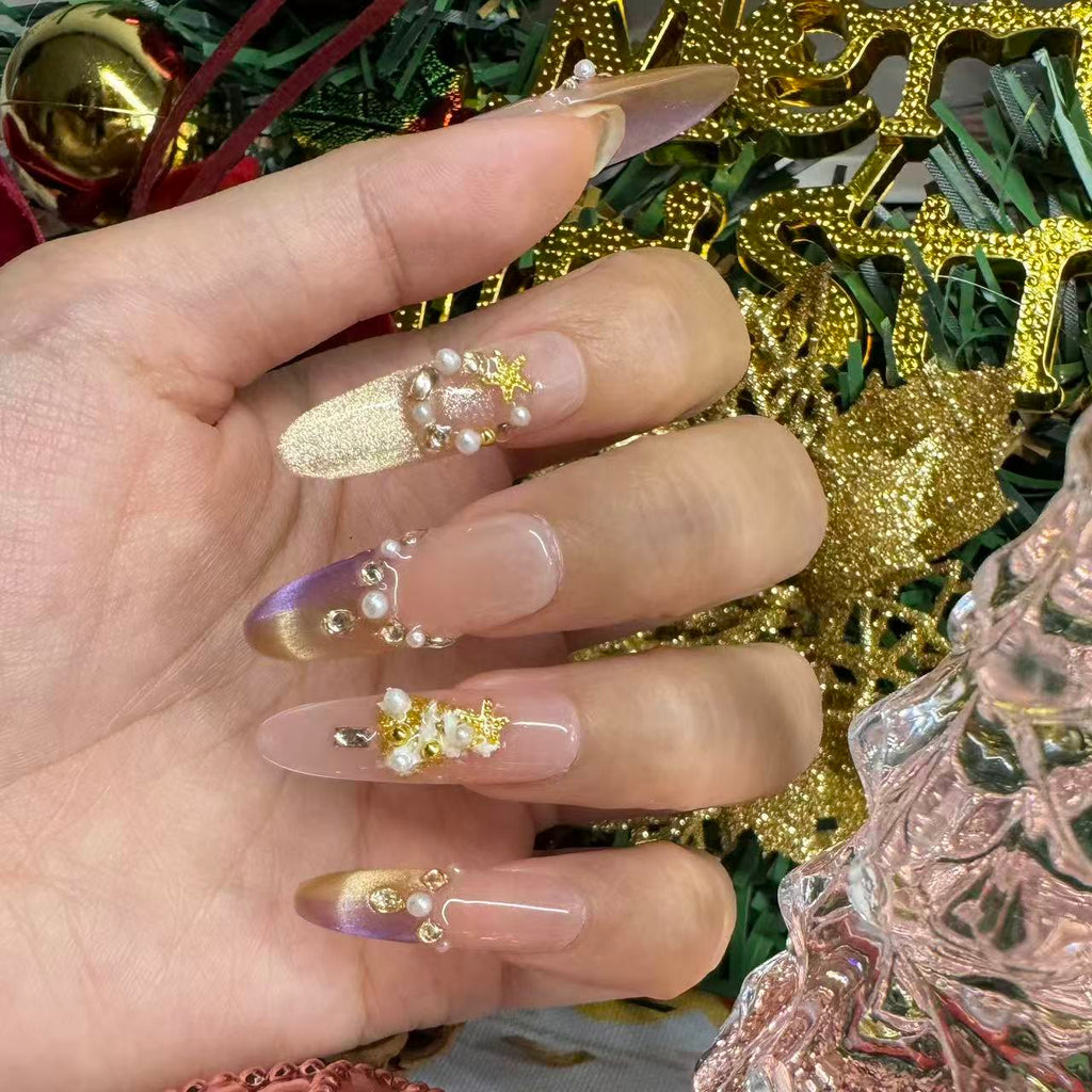 Close-up of elegant Christmas nails featuring purple-gold gradient tips, pearl clusters and gold foil details in a festive setting.