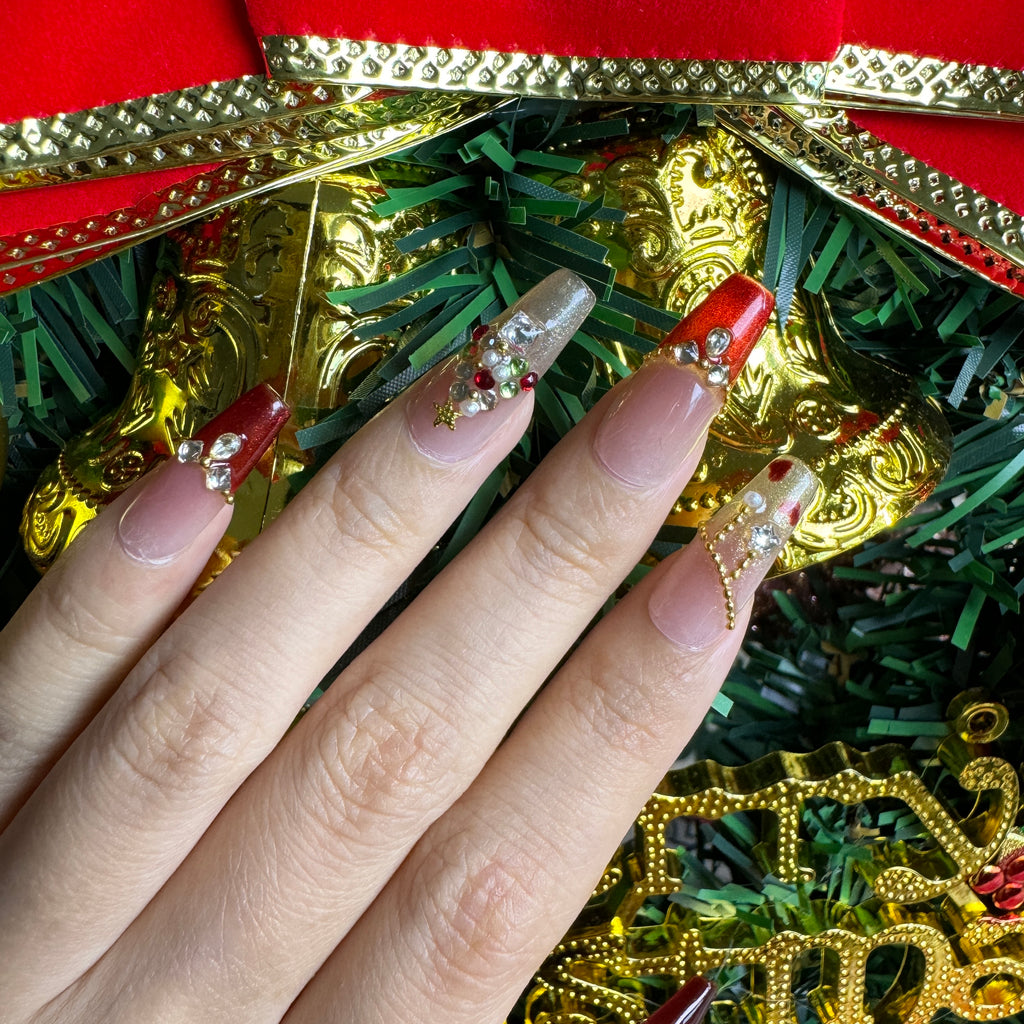 Festive red and gold Christmas nails featuring a jeweled 3D tree charm, glitter gradients and crystal details, photographed with holiday ornaments.