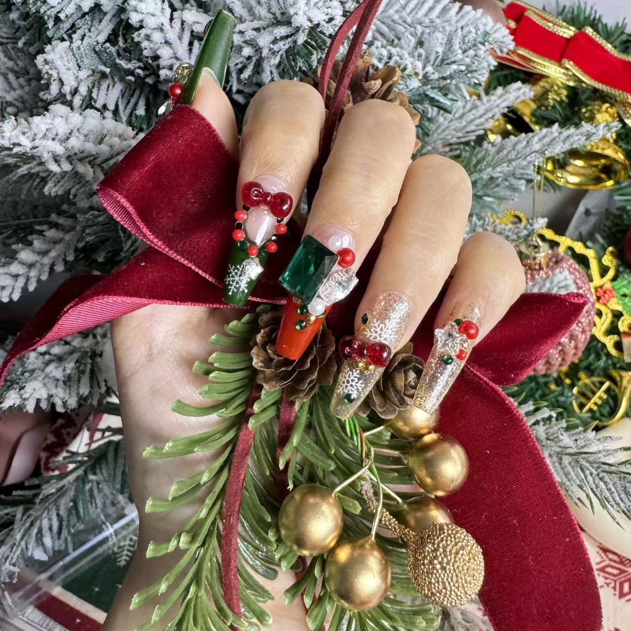 Hand wearing colorful Christmas nails featuring green gems, red ornaments, 3D bells and glitter details, photographed with festive pine branches and a red ribbon.