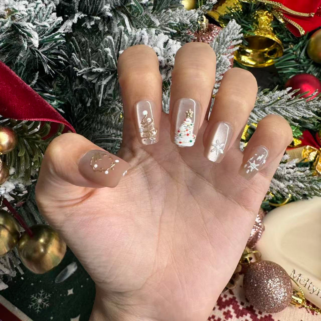 Hand with nude cat-eye Christmas press on nails with gold-painted trees, shooting stars, snow accents and a gold wreath charm arranged on Christmas holiday background.