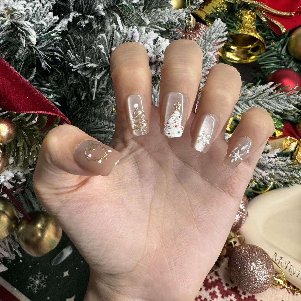 Hand with nude cat-eye Christmas press on nails with gold-painted trees, shooting stars, snow accents and a gold wreath charm arranged on Christmas holiday background.